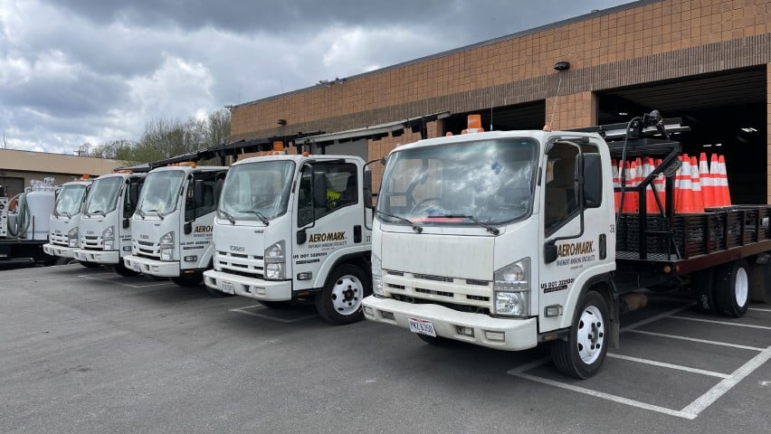 A row of white trucks with “AERO-MARK” branding on driver side door parked in front of a building, one with traffic cones on its flatbed.